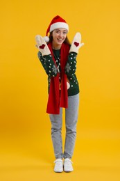 Photo of Happy young woman in Christmas sweater, Santa hat and knitted mittens on orange background