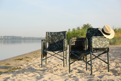 Camouflage fishing chairs with hat on sandy beach near river