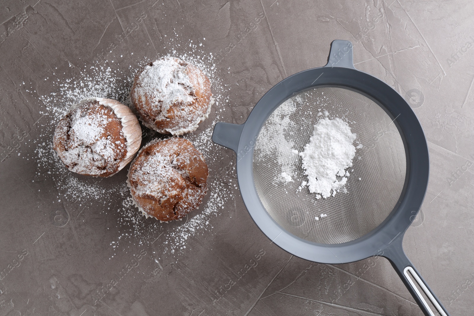 Photo of Sieve with sugar powder and muffins on grey textured table, flat lay