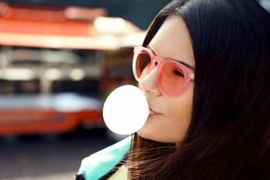 Photo of Beautiful young woman in pink glasses blowing chewing gum outdoors, closeup