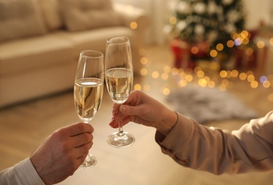 Photo of Couple clinking glasses of champagne in room decorated for Christmas, closeup. Holiday cheer and drink
