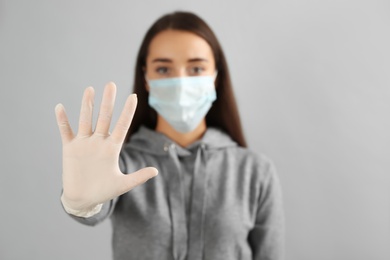 Photo of Woman in protective face mask and medical gloves showing stop gesture against grey background, focus on hand