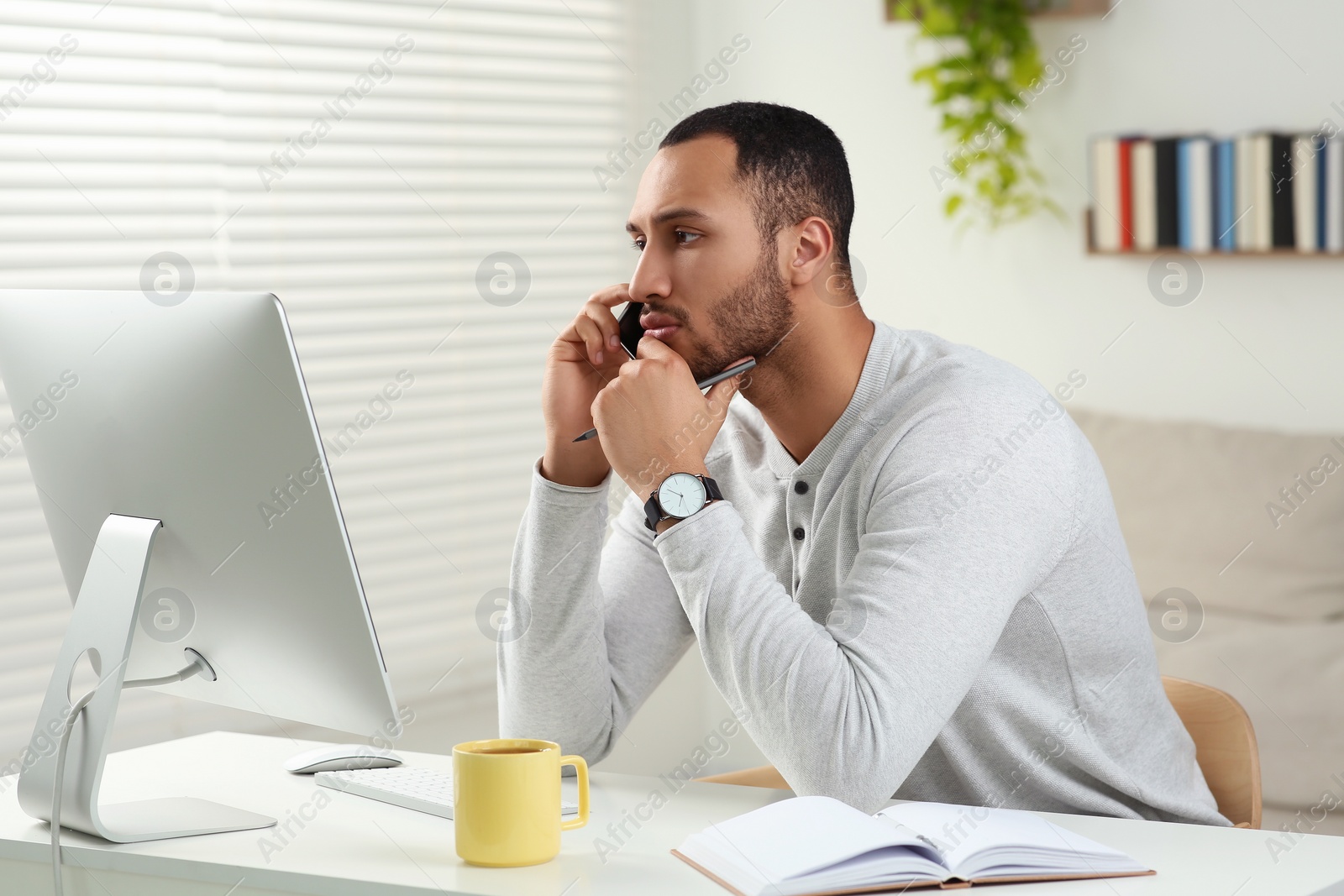 Photo of Young man talking on smartphone while working with computer at desk in room. Home office