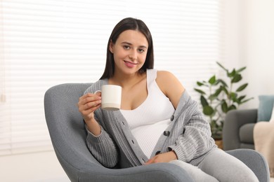 Photo of Beautiful pregnant woman with cup of drink in armchair at home