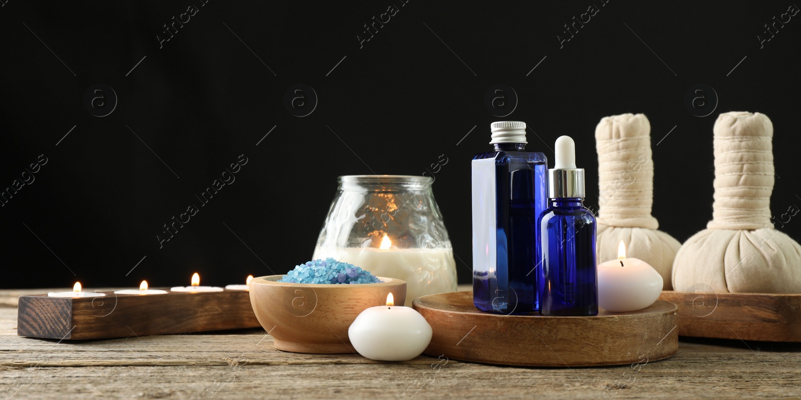 Photo of Spa composition. Cosmetic products, herbal bags, burning candles and sea salt on wooden table against black background, space for text