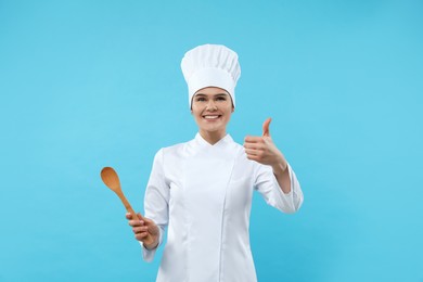 Photo of Emotional female chef with wooden spoon showing thumbs up on light blue background