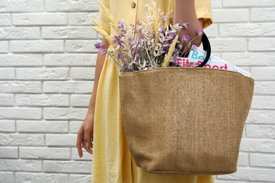 Woman holding beach bag with beautiful bouquet of wildflowers and magazines near white brick wall, closeup