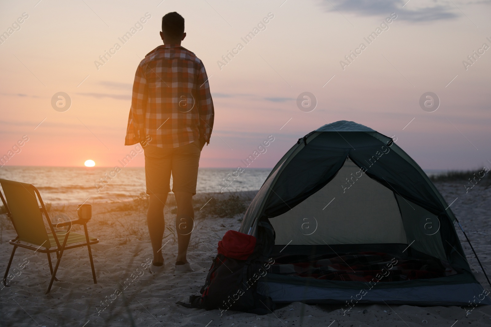 Photo of Man enjoying sunset near camping tent on beach, back view