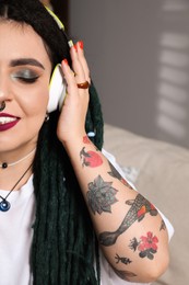 Photo of Beautiful young woman with tattoos on body, nose piercing and dreadlocks listening to music indoors, closeup