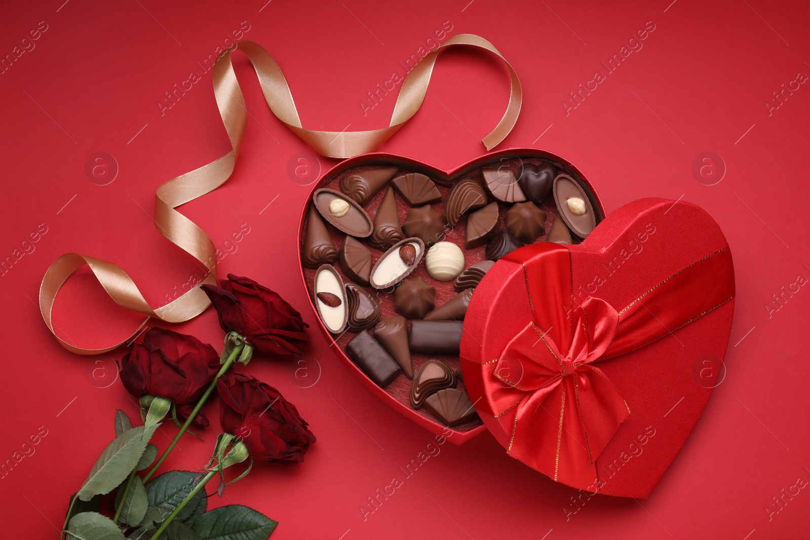 Photo of Heart shaped box with delicious chocolate candies, roses and ribbon on red background, flat lay