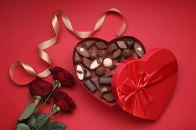 Heart shaped box with delicious chocolate candies, roses and ribbon on red background, flat lay