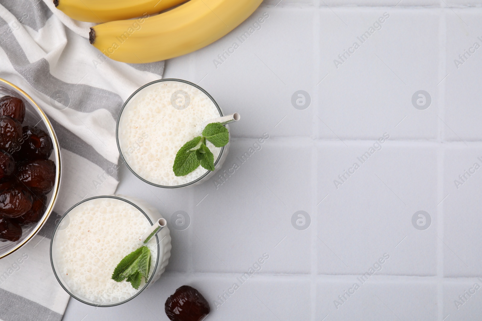 Photo of Glasses of delicious date smoothie with mint, bananas and dried fruits on white tiled table, flat lay. Space for text