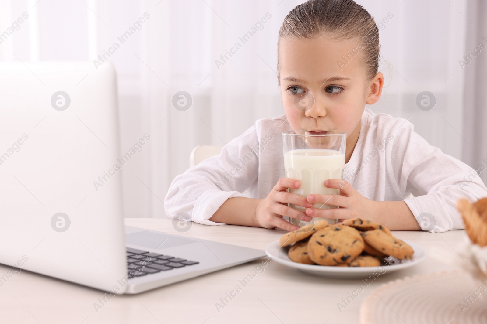 Photo of Little girl using laptop while having breakfast at table indoors. Internet addiction