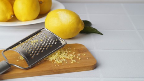 Photo of Wooden board with grater and fresh lemon zest on white tiled table, space for text