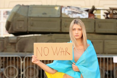 Photo of Sad woman wrapped in Ukrainian flag holding poster with words No War near broken tank on city street