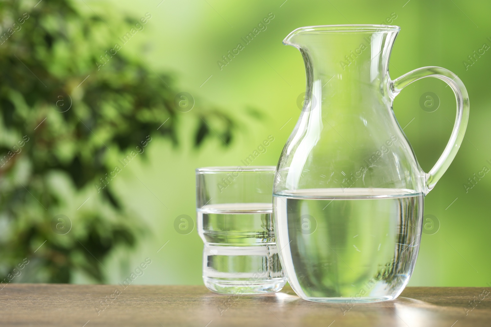 Photo of Jug and glass with clear water on wooden table against blurred green background, closeup. Space for text