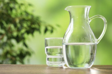 Jug and glass with clear water on wooden table against blurred green background, closeup. Space for text