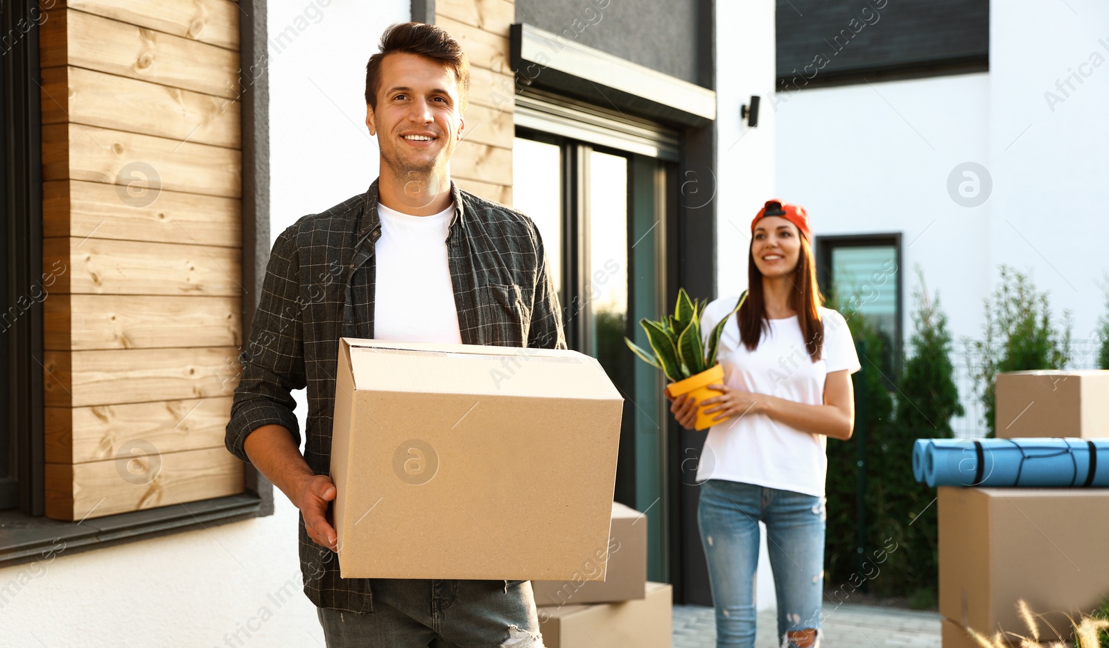 Photo of Happy couple with moving boxes and household stuff near their new house on sunny day