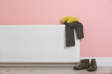 Photo of Heating radiator with knitted cap, scarf and shoes near color wall