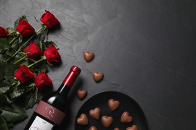Bottle of red wine, beautiful roses and heart shaped chocolate candies on black table, flat lay. Space for text