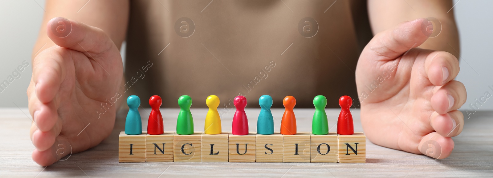 Image of Woman protecting colorful pawns and wooden cubes with word Inclusion at white wooden table, closeup. Banner design