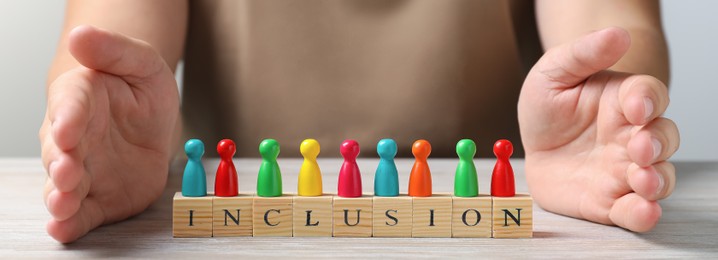 Image of Woman protecting colorful pawns and wooden cubes with word Inclusion at white wooden table, closeup. Banner design