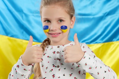 Photo of Little girl with face paint showing thumbs up near Ukrainian flag