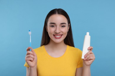 Happy young woman holding plastic toothbrush and tube of toothpaste on light blue background