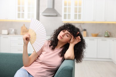 Young woman waving hand fan to cool herself on sofa at home