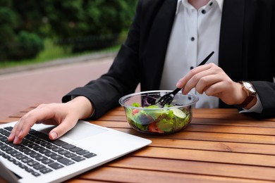 Photo of Businesswoman with container of salad using laptop while having lunch at wooden table outdoors, closeup