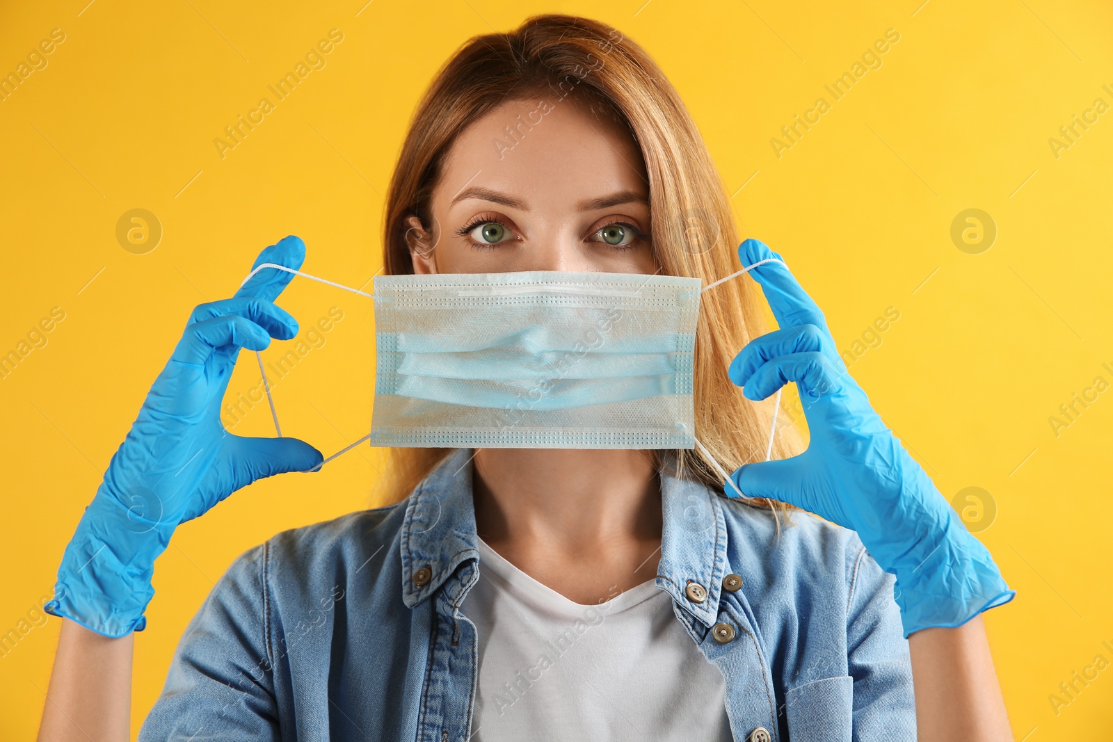 Photo of Young woman in medical gloves putting on protective face mask against yellow background