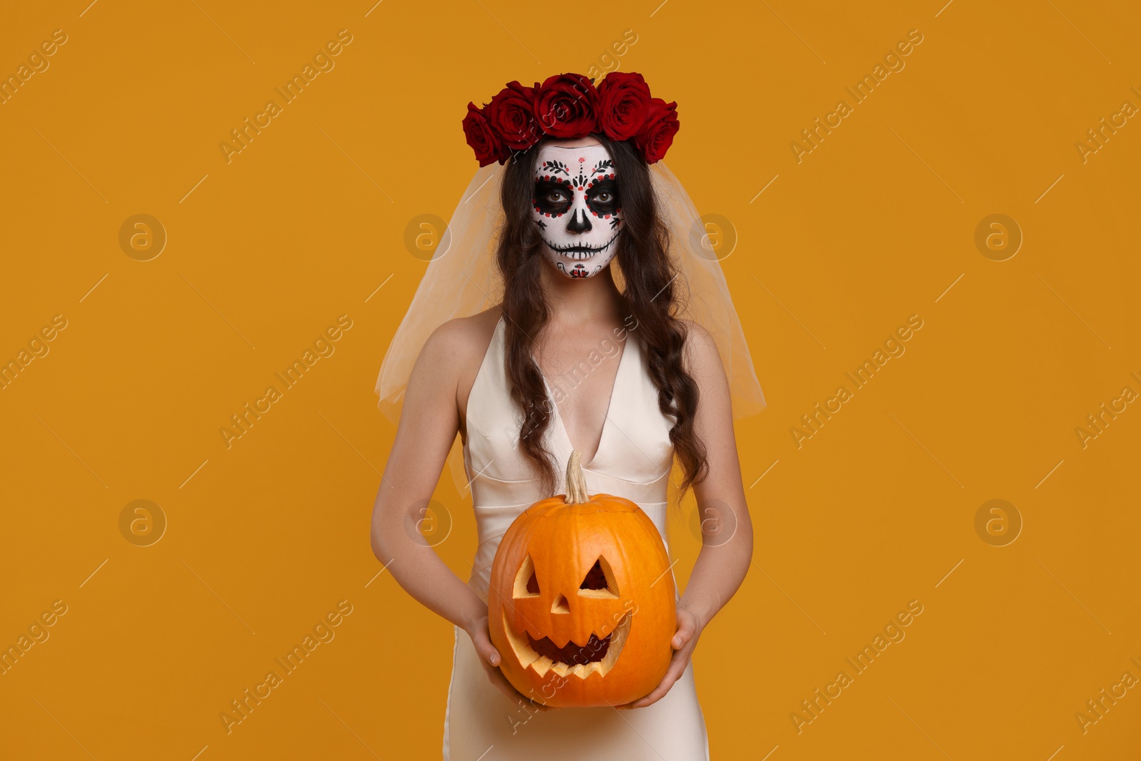 Photo of Young woman in scary bride costume with sugar skull makeup, flower crown and carved pumpkin on orange background. Halloween celebration