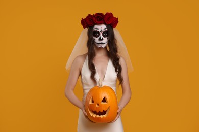 Young woman in scary bride costume with sugar skull makeup, flower crown and carved pumpkin on orange background. Halloween celebration