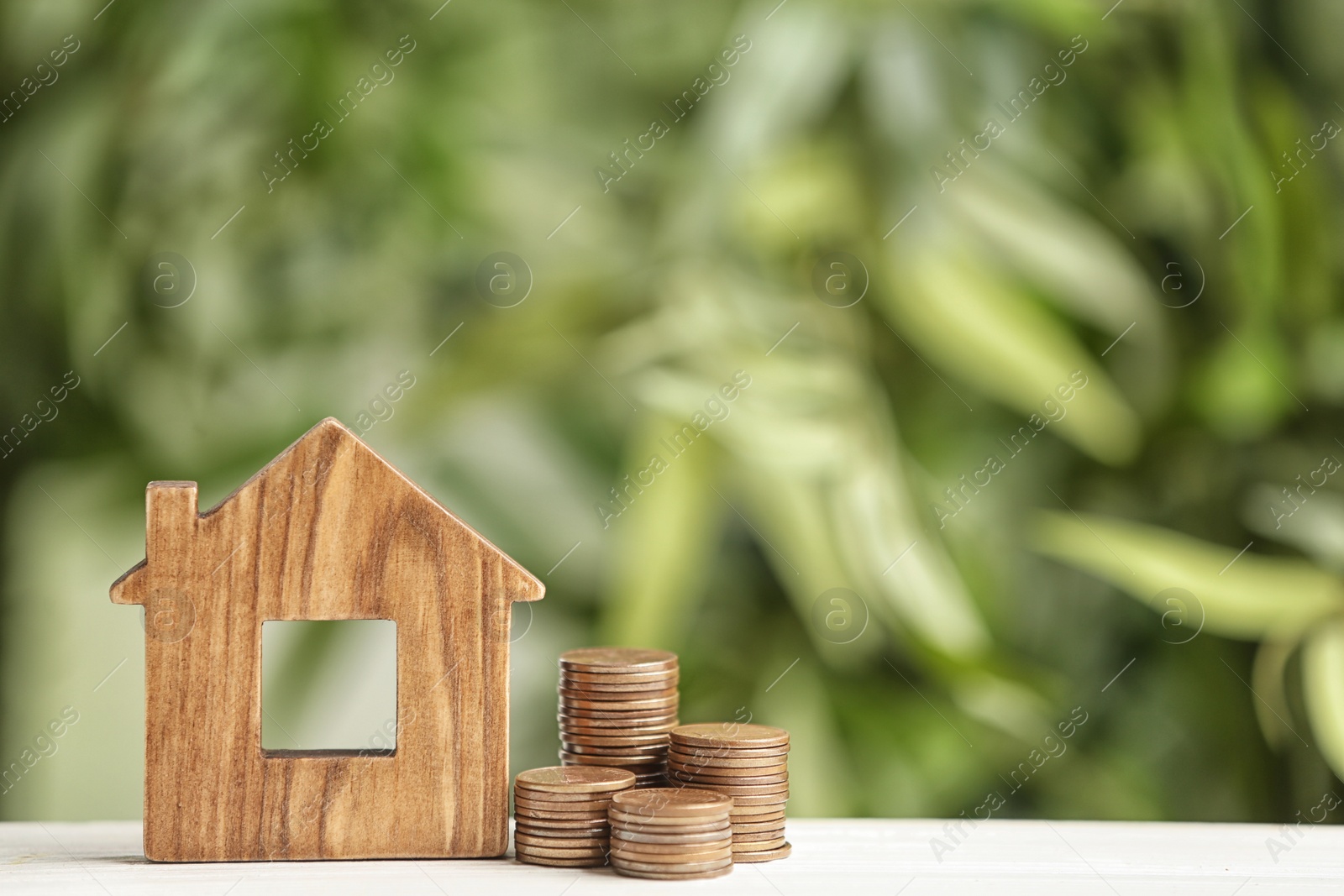 Photo of House model and coins on white table against blurred green background. Space for text