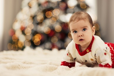 Photo of Cute little baby in Christmas sweater on knitted blanket against blurred festive lights, space for text. Winter holiday