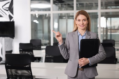 Happy real estate agent with leather portfolio pointing indoors. Space for text