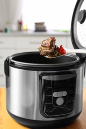 Photo of Spoon with meat and vegetables over modern multi cooker on table in kitchen