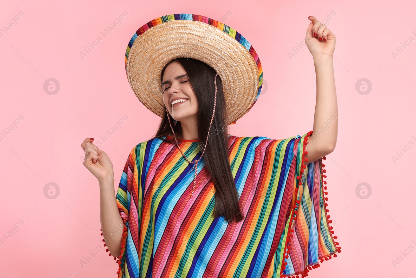 Photo of Young woman in Mexican sombrero hat and poncho dancing on pink background