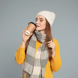 Happy young woman in warm clothes with drink on grey background. Winter season