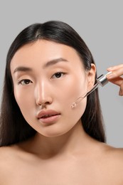 Beautiful young woman applying cosmetic serum onto her face on grey background