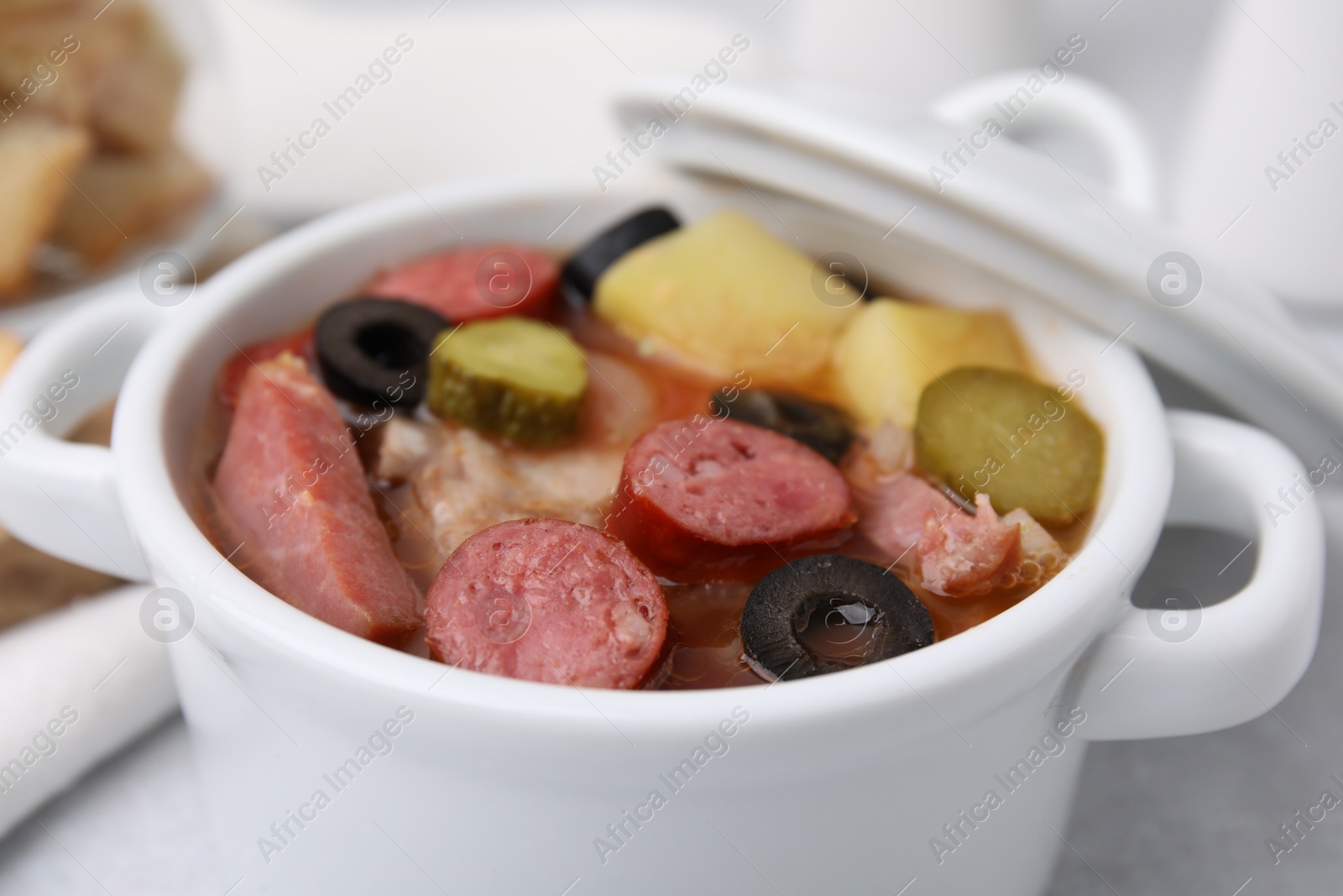 Photo of Meat solyanka soup with thin dry smoked sausages in pot on table, closeup