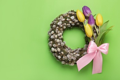 Wreath made of beautiful willow, colorful tulip flowers and pink bow on green background, top view. Space for text