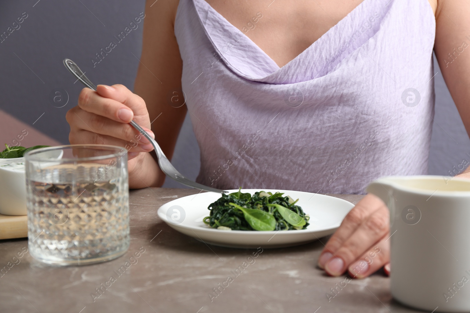 Photo of Young woman eating tasty cooked spinach at grey marble table, closeup. Healthy food