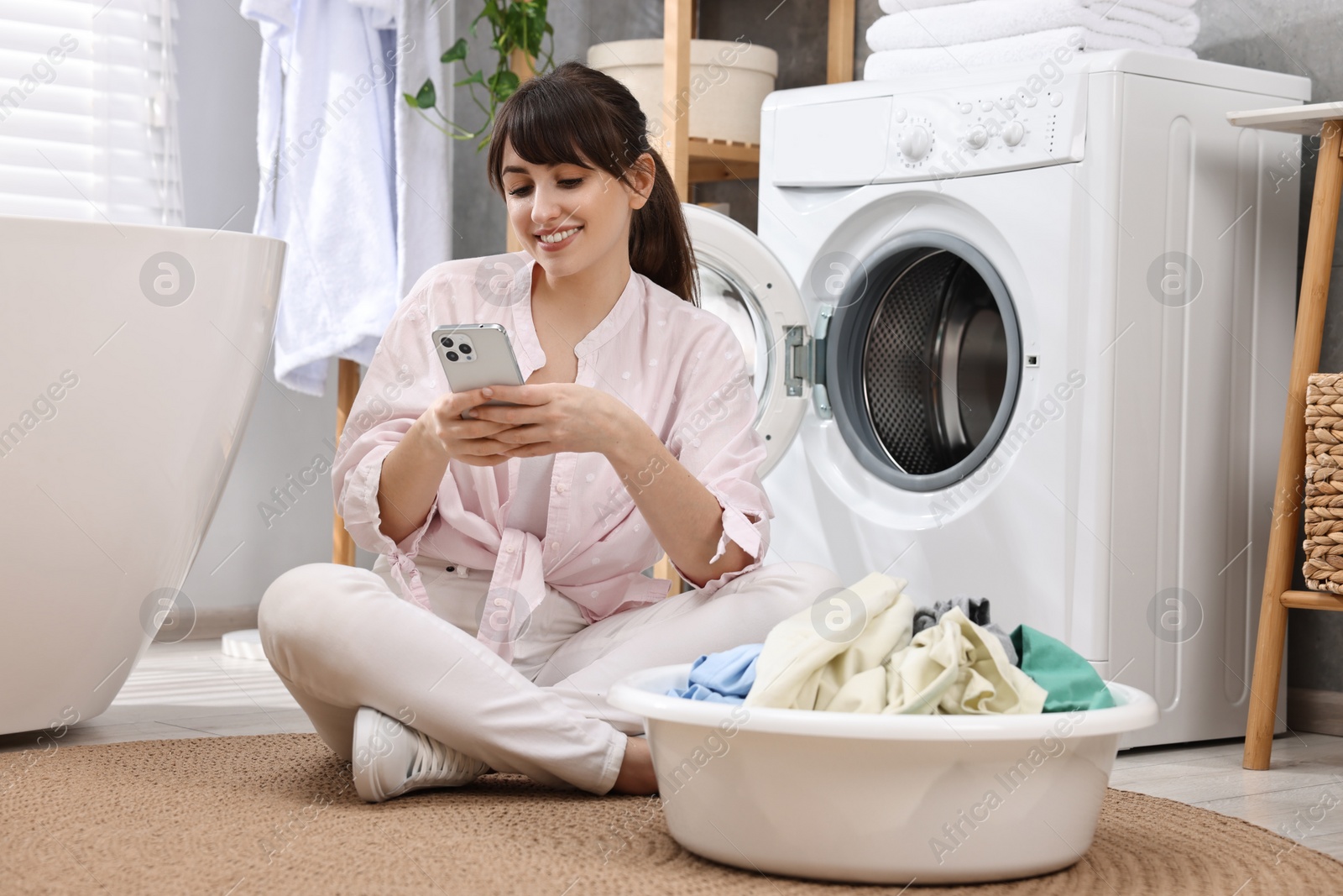 Photo of Happy young housewife with laundry using smartphone near washing machine at home