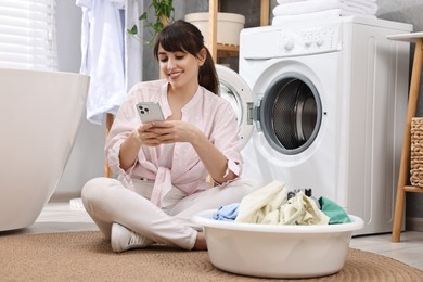 Happy young housewife with laundry using smartphone near washing machine at home