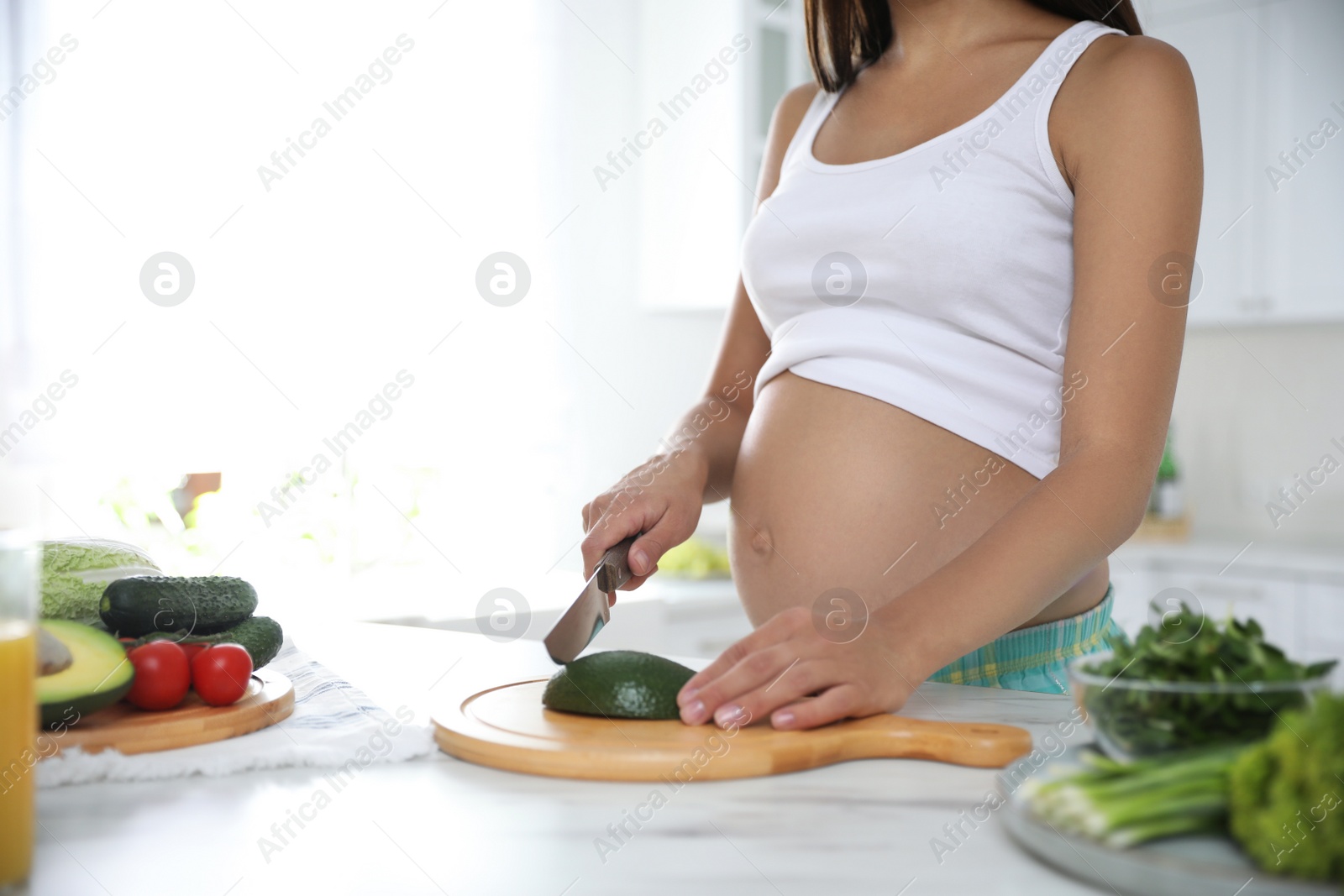 Photo of Young pregnant woman cutting avocado at table in kitchen, closeup. Taking care of baby health