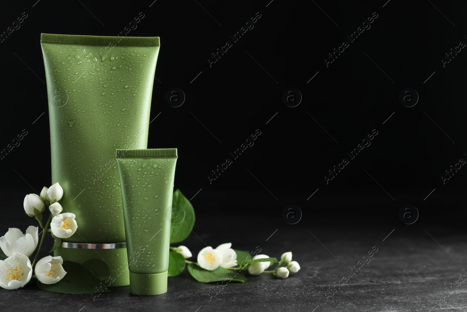 Photo of Tubes of cosmetic products and flowers on grey stone table against black background. Space for text