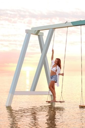 Young woman enjoying sunrise on swing over water