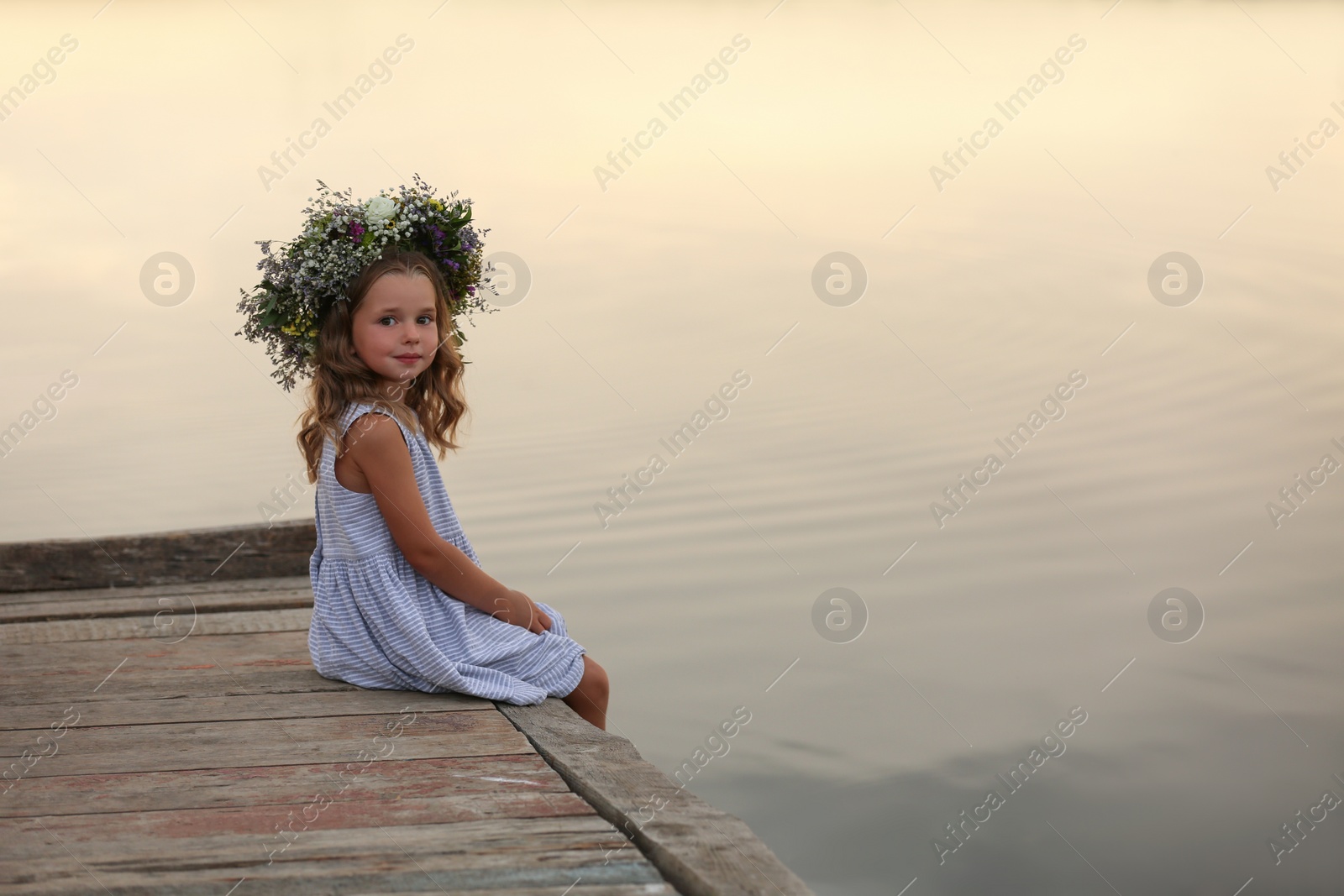 Photo of Cute little girl wearing wreath made of beautiful flowers on pier near river