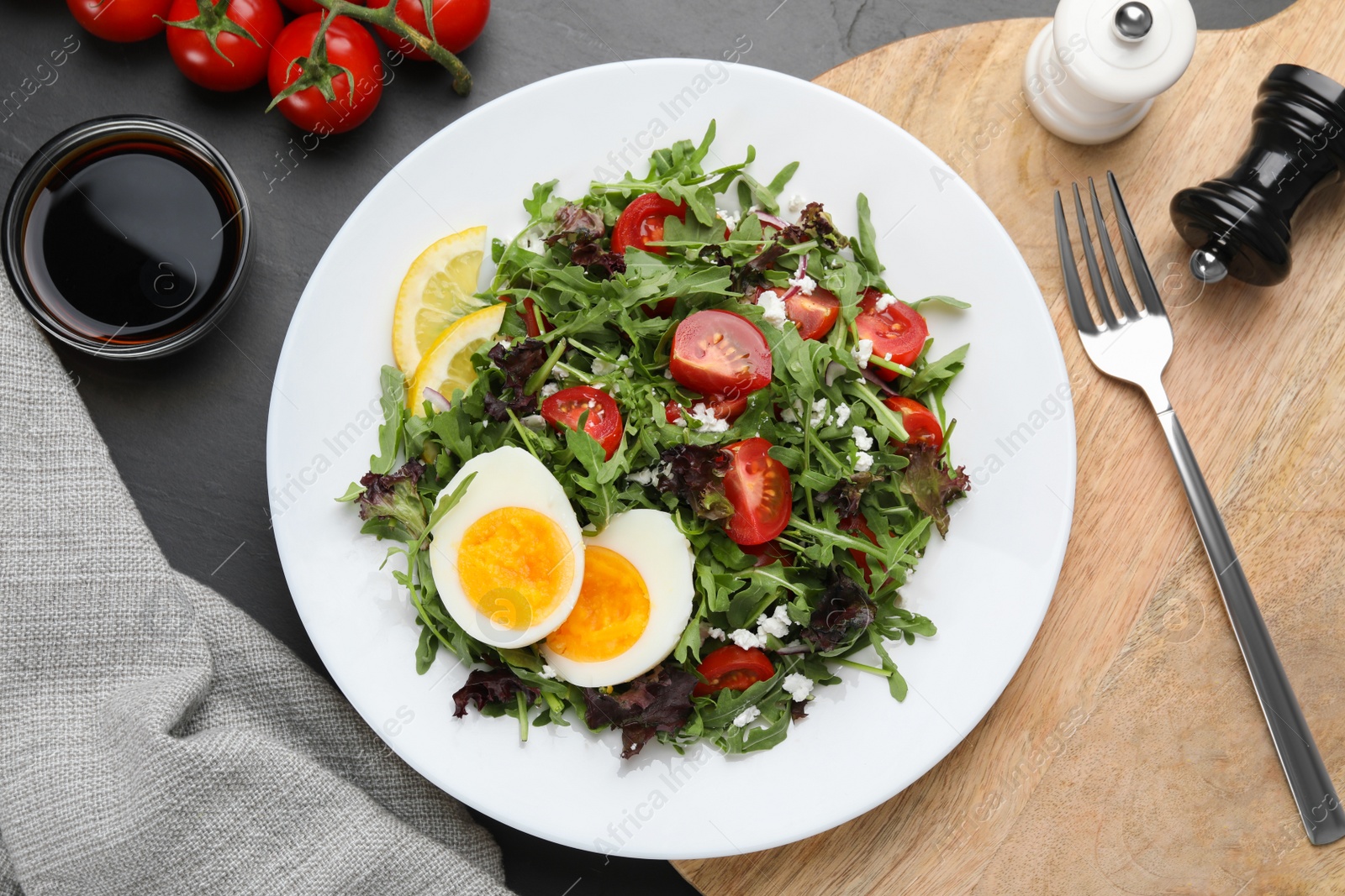 Photo of Delicious salad with boiled egg, arugula and tomatoes served on black table, flat lay